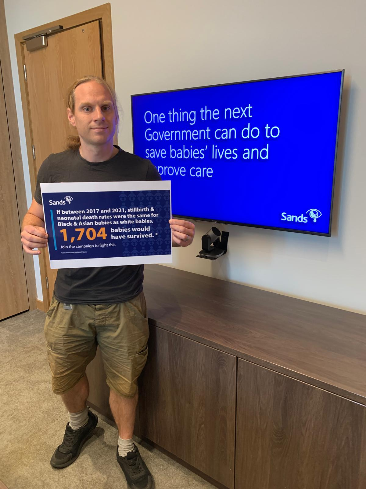 A person holding a sign that reads "If between 2017 and 2021, stillbirth and neonatal death rates were the same for Black & Asian babies as white babies, 1,704 babies would have survived".