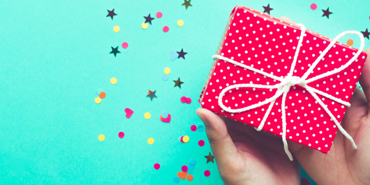 a small gift, wrapped in red paper tied with string, is held over a turquoise background with heart, star and circle shaped confetti 