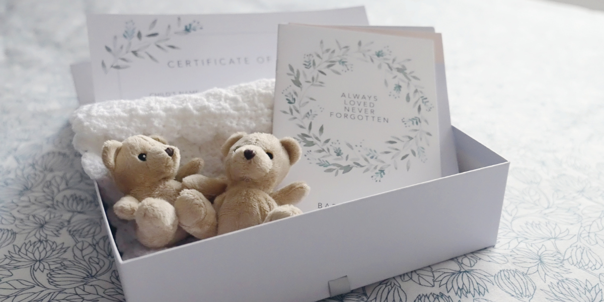 A white box with 2 small teddy bears placed on a white knitted blanket, and a card reading "always loved, never forgotten"