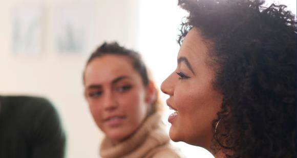 Woman with curly hair and hoop earrings speaks in a room, another woman sat by her listens