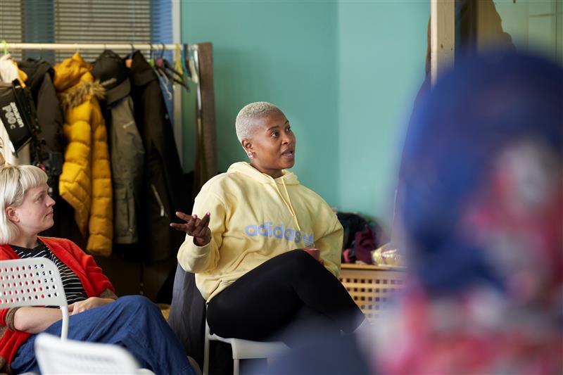 Black woman in yellow hoodie is sat in group, speaking. White woman in red cardigan is sat next to her listening.