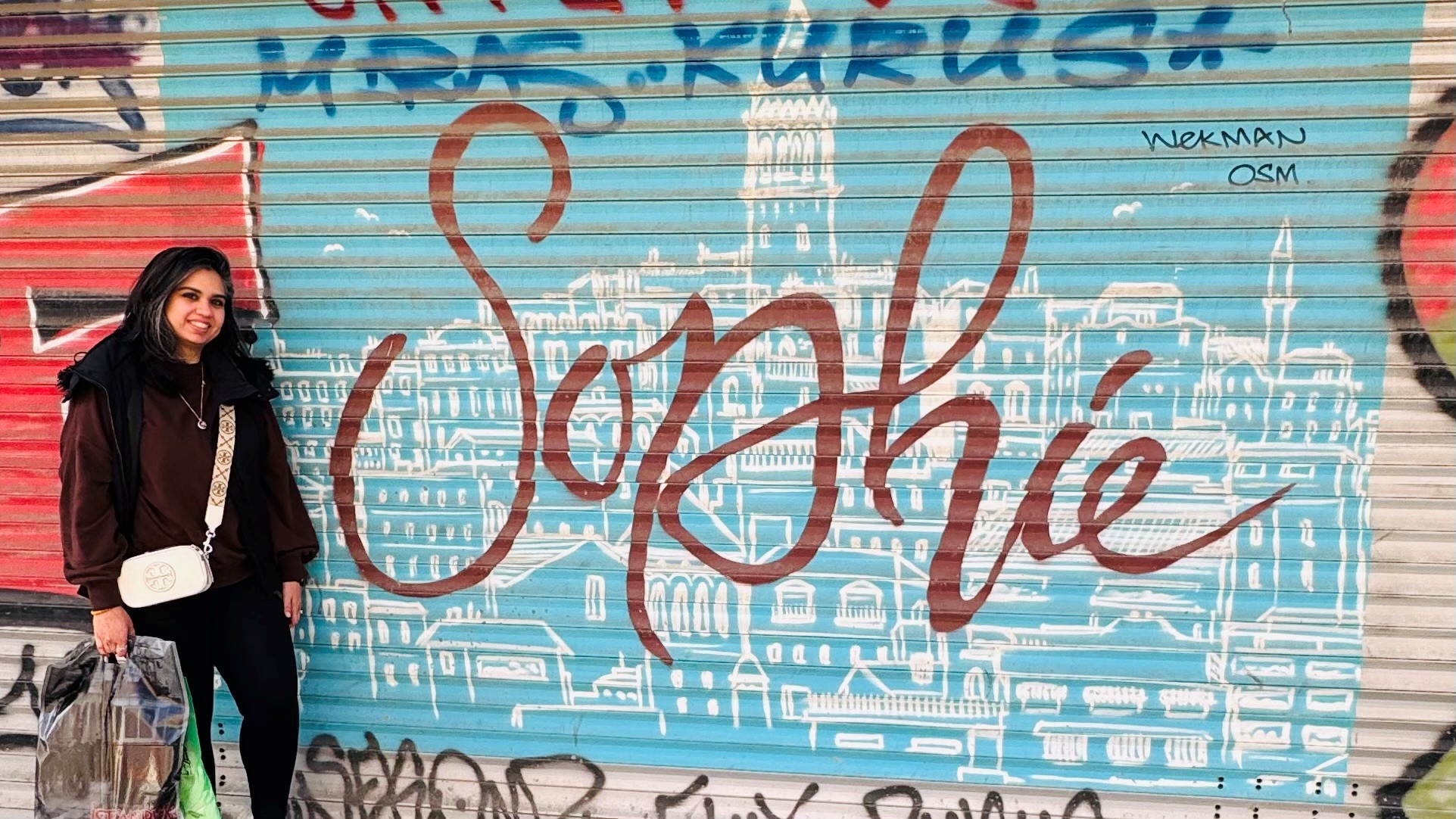 A South Asian woman called Naveen stands next to a metal shop front shutter that has the name Sophia spray painted on it in large cursive letters. Naveen is wearing a black coat and carrying a white bag.