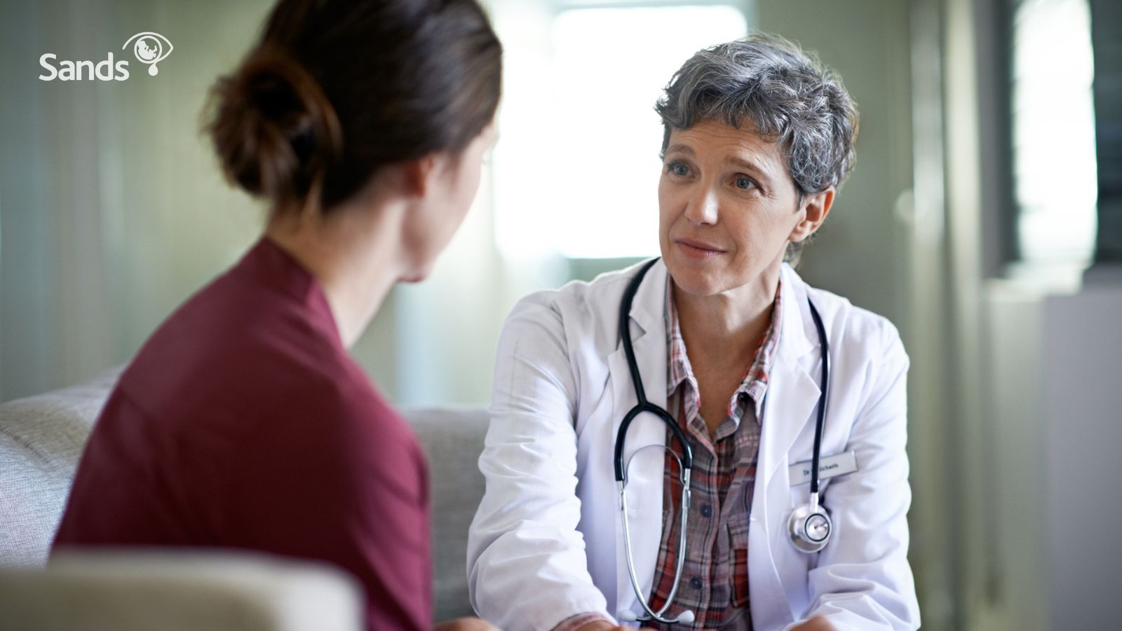 A female healthcare worker with short grey hair wearing a white coat with a stethoscope around her neck looks with an expression of understanding toward a women with her back to the camera