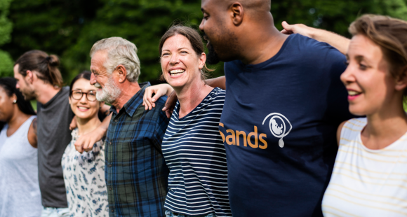 People of different ages and ethnic backgrounds standing in a line, hugging, smiling at each other, wearing Sands t-shirts