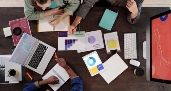 A group of people gathered around a table with laptops and notepads, having a discussion.