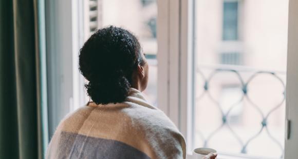 Lady sat with a blanket wrapped around her looking out of the window holding a cup of tea