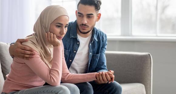 A man wearing a blue shirt has his arm around a lady wearing a headscarf as they are sat on the sofa