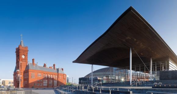 A picture showing two buildings, one redbrick with a clock tower, the other a modern looking building with a long roof and columns.
