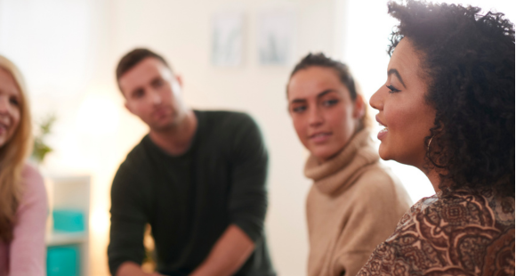 A woman with dark, shoulder length, curly hair is pictured from her left side speaking to a group. Two onlookers, a man and a woman look and listen