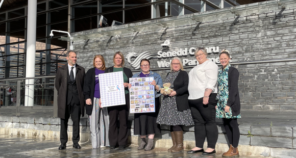 A group of people stood outside a large building made of glass and concrete. They are holding signs, and the building says "Senedd Cymru Welsh Parliament" on it.