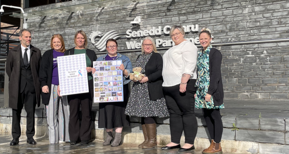 A group of people standing outside a large building made of concrete and glass. The people are holding a signs. The building says "Senedd Cymru Welsh Parliament" on it.