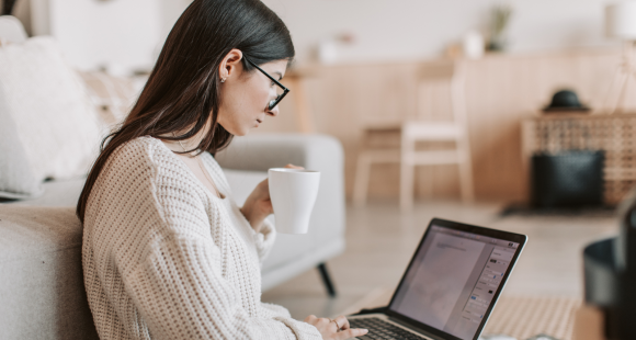 Woman sitting on the floor with her back to a sofa. She is using her laptop on her lap and holding a mug of tea