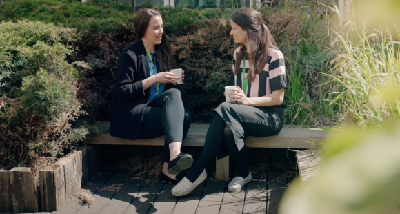 Two women sitting on a bench and talking, both are holding cups of tea.