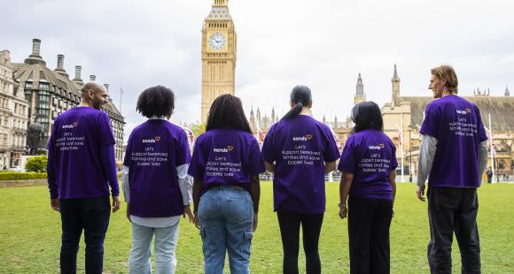A group of people standing outside the UK Parliament with big ben in the background wearing purple T-Shirts which says 'Sands: Let's support bereaved families and save babies' lives'