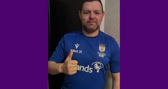 A man smiling at the camera and giving a thumbs up wearing a blue Sands United football shirt with the name Ellie on after his daughter who was stillborn after a Termination for Medical Reasons (TFMR)