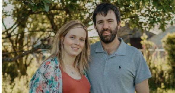 A lady with long blonde hair and an orange dress smiling at the camera with a man with dark hair with his arm around her wearing a gret t-shirt smiling at the camera with a background of trees