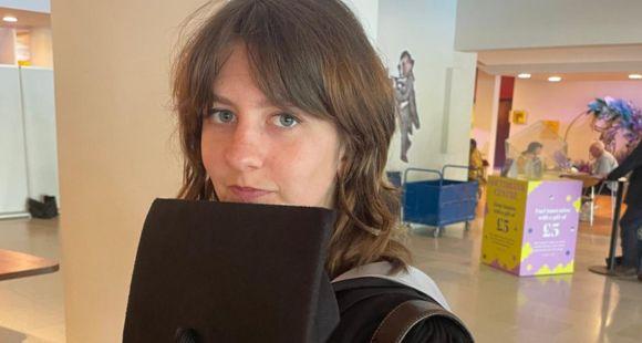 Young lady with brown hair holding a black graduation cap looks at the camera as she explains how she made a documentary after her brother was stillborn when she was six.
