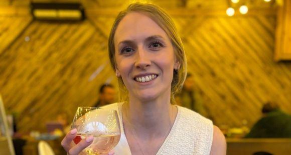 A lady with blonde hair holding a glass of wine and smiling at the camera