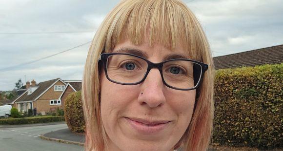 A lady with blonde hair and a fringe and black glasses smiling at the camera