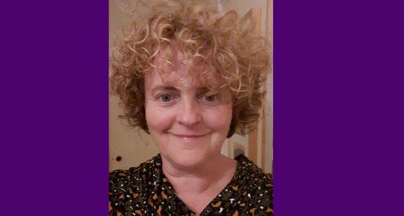 A lady with blonde curly hair smiles at the camera after sharing her self penned poem about stillbirth.