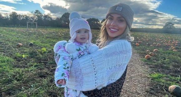 A lady wearing a grey bobble hat holding a baby girl in a hat and a snowsuit and smiling at the camera