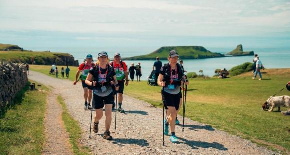A group of people walking on a gravel path, in the background there is green grass, dog walkers and the irish sea.