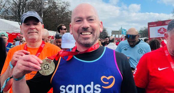 A man wearing a purple Sands running vest holding a gold medal up and smiling at the camera