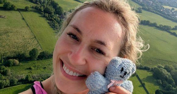 A lady at the camera with blonde hair smiles whilst holding a knitted blue teddy that belonged to her son who was a stillbirth.