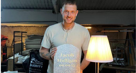 Man in a grey t-shirt smiling at the camera and holding a grey small headstone that reads Jacob Mulligan in gold letters 09.09.15