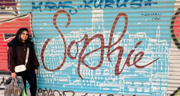 A South Asian woman called Naveen stands next to a metal shop front shutter that has the name Sophia spray painted on it in large cursive letters. Naveen is wearing a black coat and carrying a white bag.