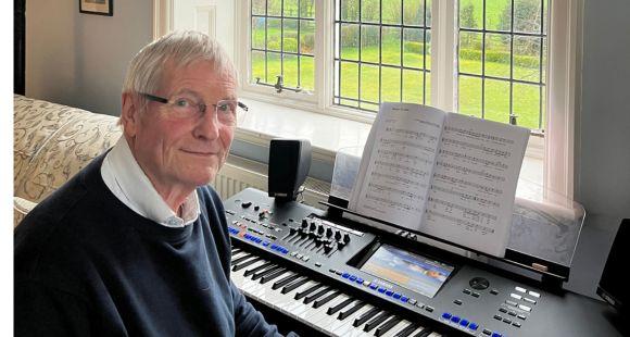 A man with glasses sat at a keyboard piano with open music on a music stand