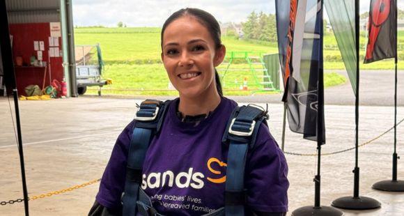 A lady with brown hair tied back wearing a Sands purple t-shirt and a harness smiles at the camera after speaking about the impact of Sands memory boxes.