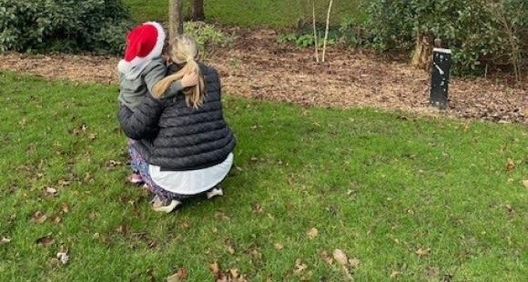 A child in a Santa hat cuddling a lady with  blonde hair in a ponytail wearing a black jacket looking at a tree on grassland