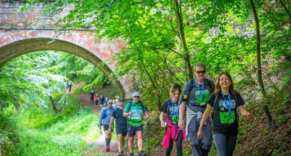 A long line of people walking on a path, in the background there are trees with green leaves on a hill and an orange brick bridge.