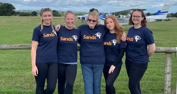 five women all wearing Sands t-shirts in front of a plane