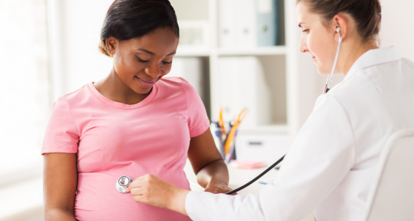 A female doctor in a white coat uses a stethoscope on the stomach of a pregnant Black woman wearing a pink top