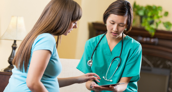 A woman wearing a light green t-shirt gestures toward a tablet device held by a female healthcare worker that they are both looking at 