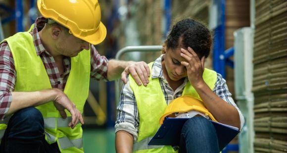A white male warehouse worker in a red check shirt, hi-vis jacket and yellow hard hat puts their hand comfortingly on the shoulder of a Black female co-worker wearing a blue check shirt and hi -via jacket. She is sitting with one hand on her forehead and looks upset.