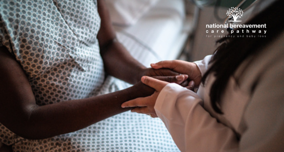 Black woman in hospital gown is sat on a bed, her hands held by a person with long hair and brown skin who is standing by her.