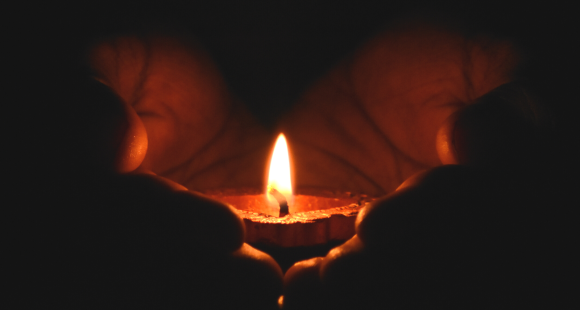 Two hands holding a diya candle, a close zoom in on the candle, very dark background 