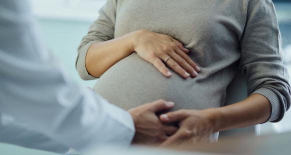 The arm of a health care worker in a white lab coat holds the left hand of a pregnant woman with her right hand on her stomach 