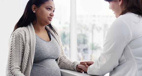 A pregnant woman wearing a white cardigan and grey t shirt is reassured by a female health worker in a white lab coat