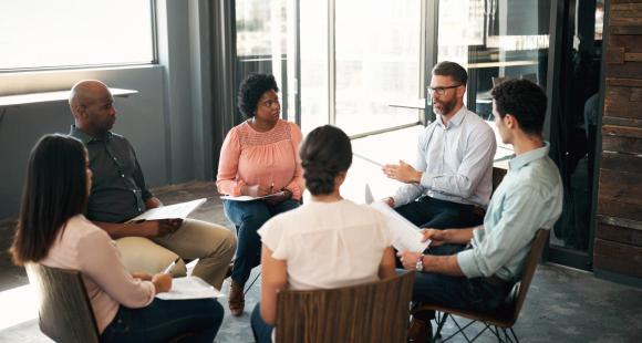 A group of men and women of mixed ethnicities sit on wooden chairs in a circle talking in a semi-formal setting