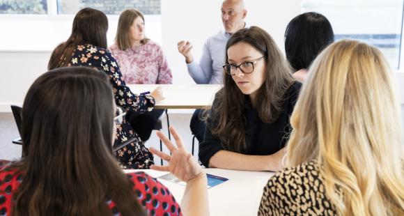 A group of women sit talking around a table. Two have their backs to the camera and the one on the left is gesturing with her hand to make a point. In the background we see another table discussion between two women and a man.