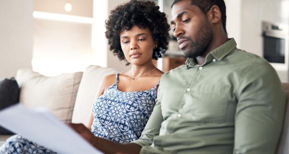 Black woman with afro and patterned blue top sits on sofa next to Black man with short hair and a green shirt, both looking at a piece of paper.