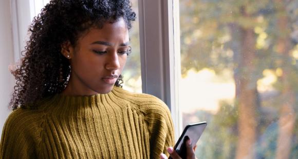 A Black woman wearing a yellow jumper looks at her mobile phone with an expression of concern