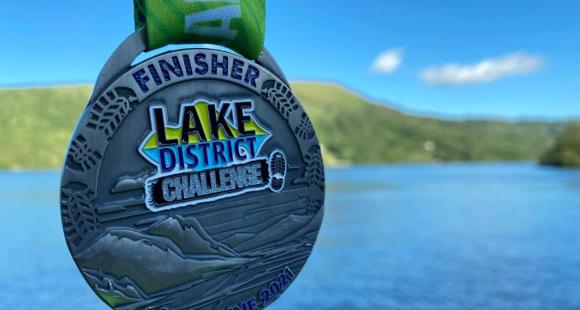 A medal with green ribbon, lsilver plate with the writing finisher medal Lake District Challenge, with Lake and green trees in the background.