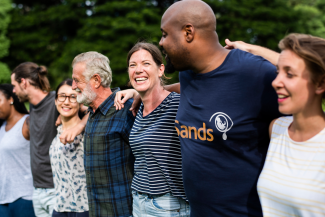 a group of people of all ages and races standing together holding each others shoulders, smiling, while wearing Sands t-shirts