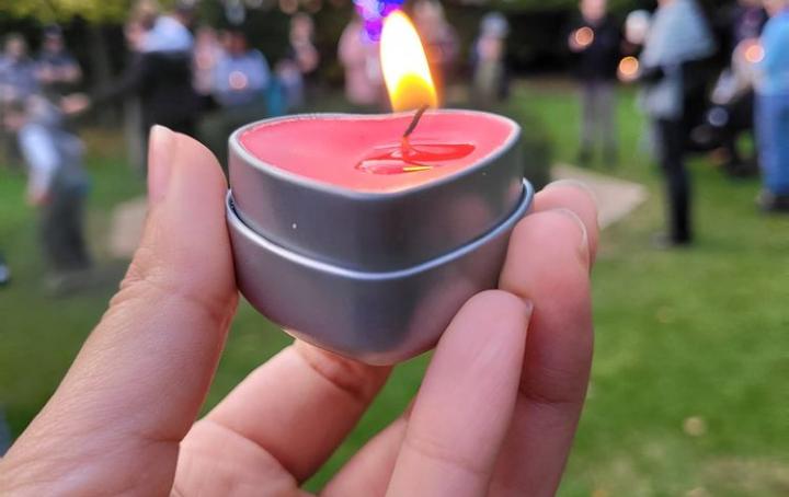 a hand holding red heart shaped candle that is lit up in a park, in the background there is a baby loss memory tree covered in pink and blue ribbons with babies' names