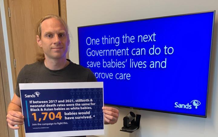 A man holding a sign that reads "if between 2017 and 2021, stillbirth & neonatal death rates were the same for Black and Asian babies as white babies, 1,704 babies would have survived."